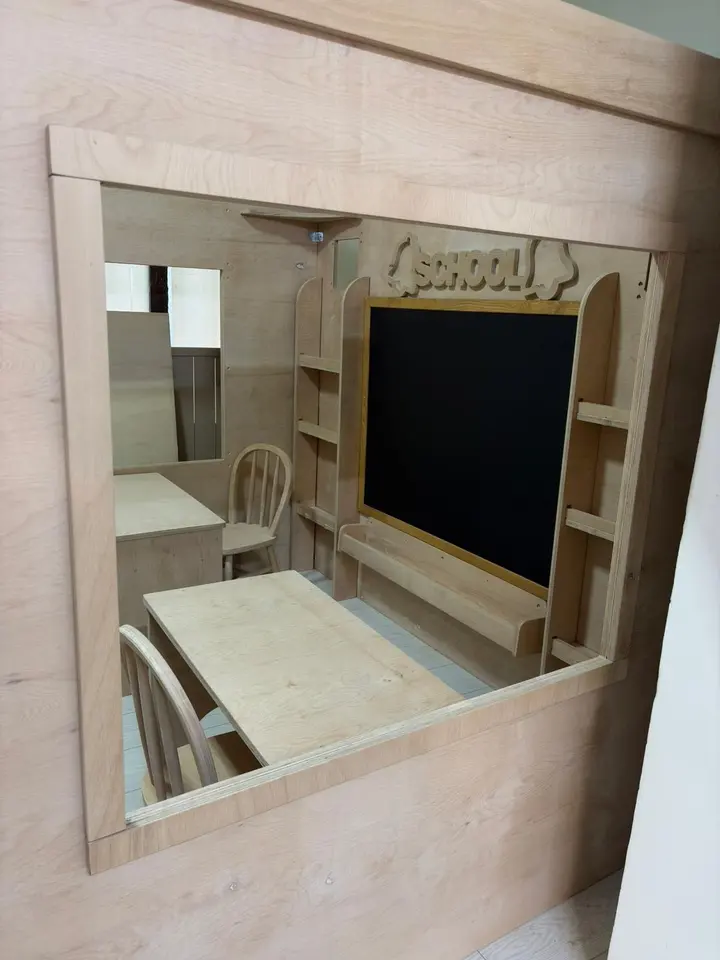Side window view into wooden school playhouse interior with desks and chalkboard